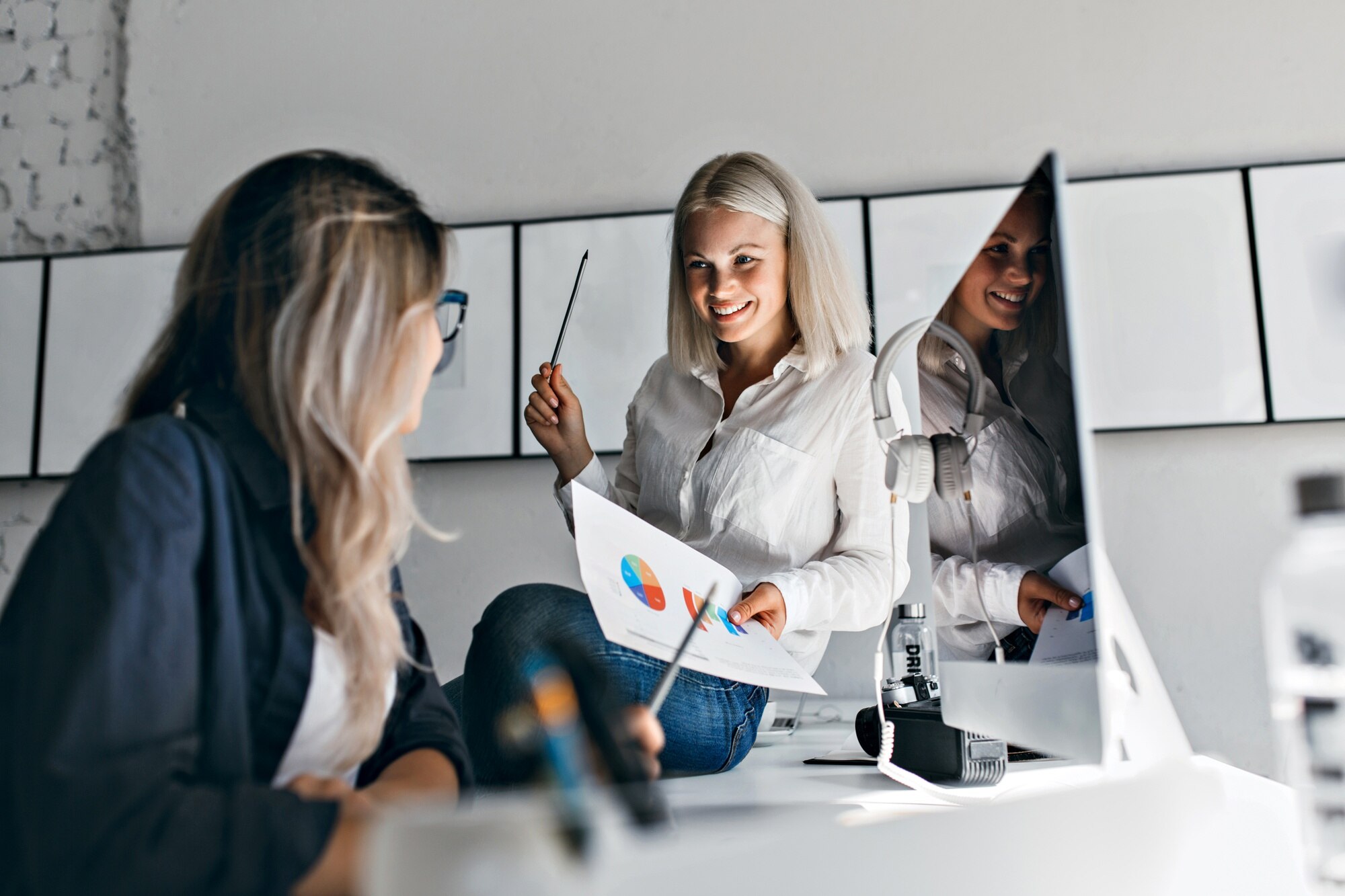 Lachende blonde vrouwelijke manager houdt infographic en potlood vast terwijl ze aan een tafel zit, met twee vrouwen die aan een computer werken in een kantoor.